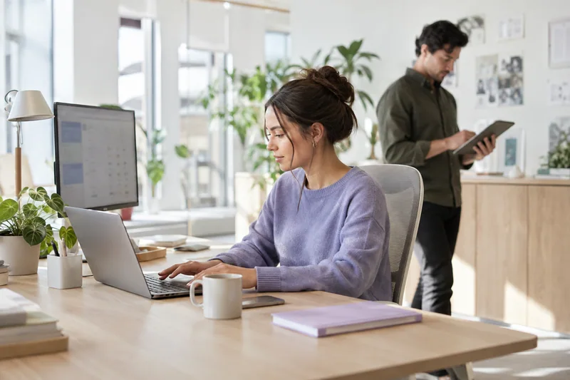 A recruitment-office coordinator using a laptop beside an external monitor, colleague checking a task on a tablet, authentic uncluttered desktop with ordinary equipment, daylight and a subtle lilac notebook. AI-generated workplace illustration.